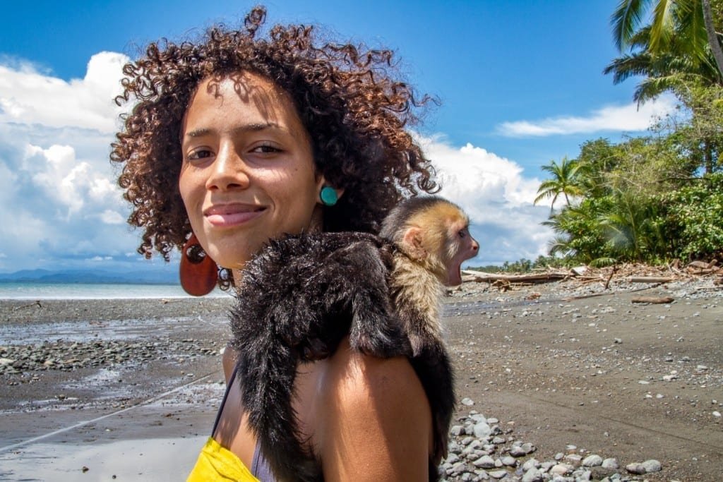 Costa-Rica-Custom-Private-Travel-Design-South-Pacific-Beach-Girl-Capuchin Girl in yellow shirt standing on a brown sand beach in the South Pacific of Costa Rica, with a small monkey clinging to her shoulder