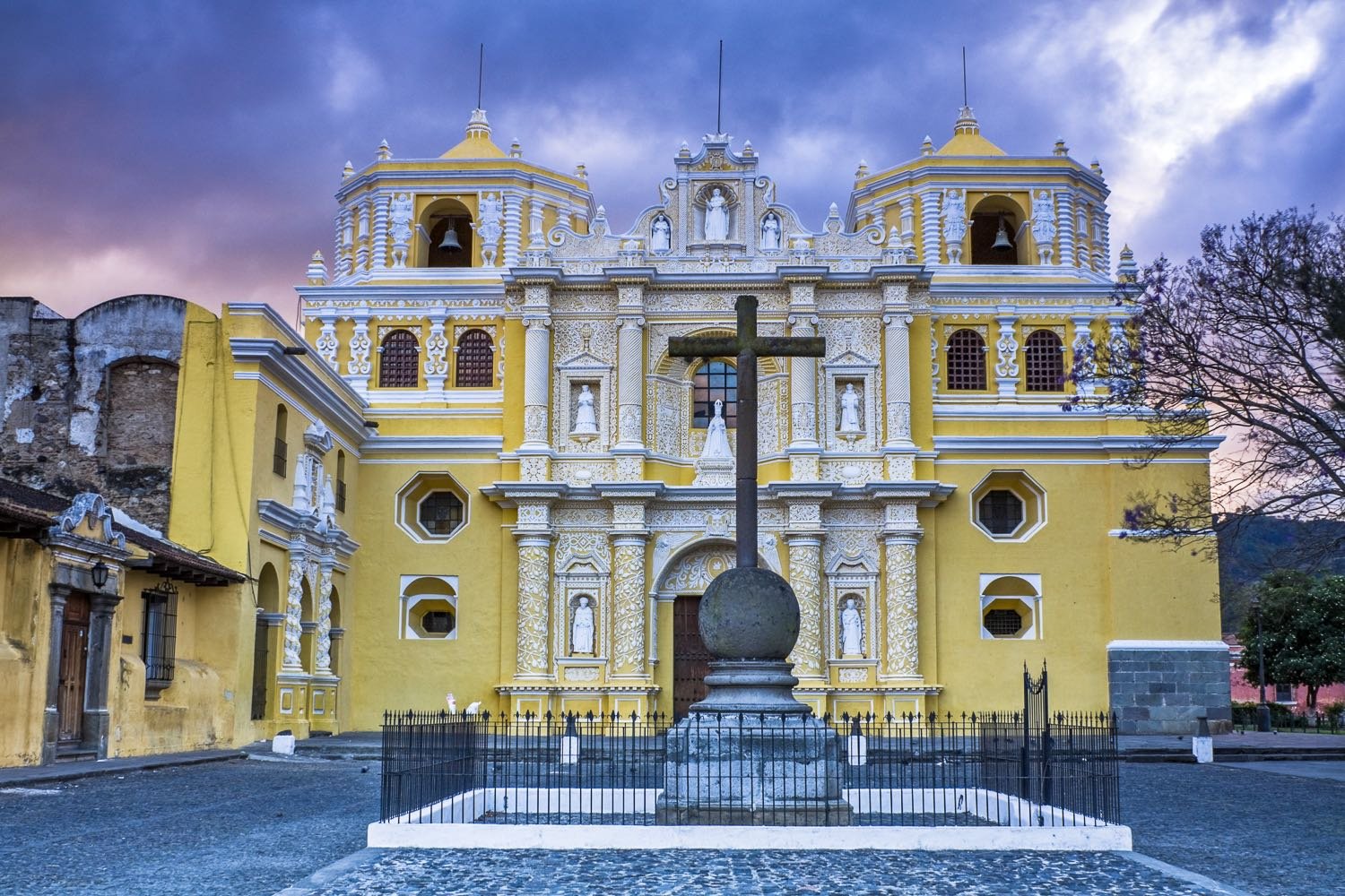 Guatemala-Private-Custom-Travel-Design-Antigua-Merced-3 Large beige and yellow La Merced Church in Antigua Guatemala with a large cross in the center of the courtyard, photographed on private travel in Guatemala