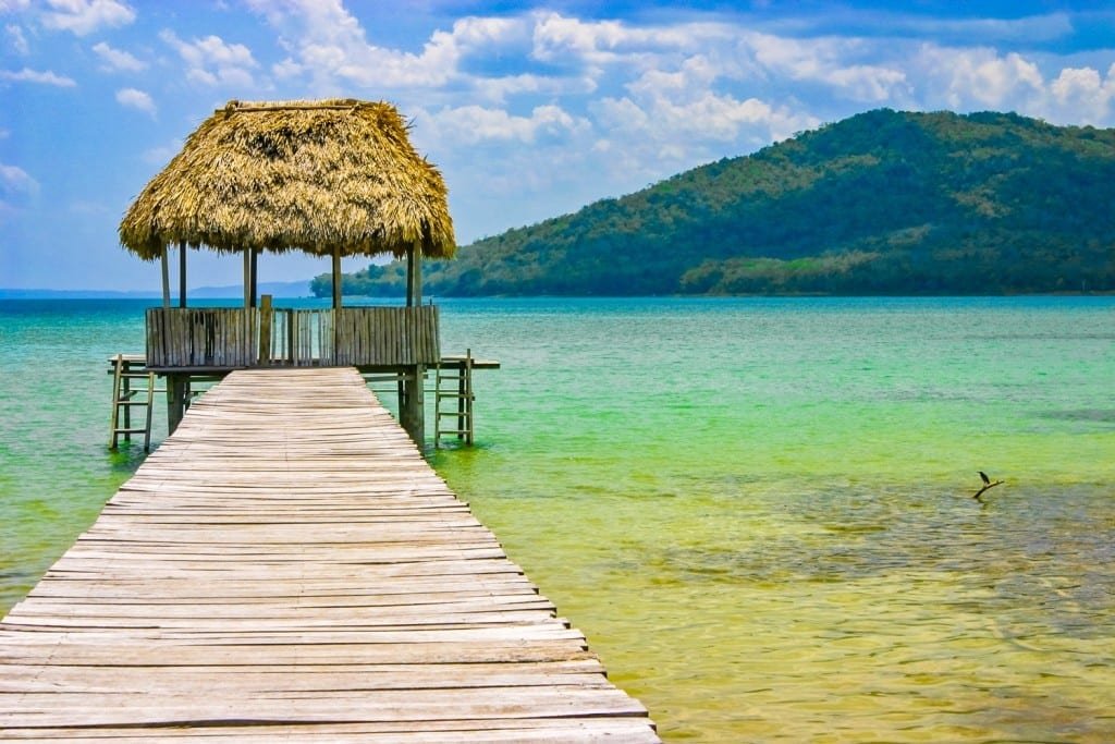 Guatemala-Private-Custom-Travel-Design-Tikal-Peten-Itza A straw roof over water bungalow on the end of a dock overlooking the Caribbean blue ocean with a volcano in the background. Peten Itza, Guatemala