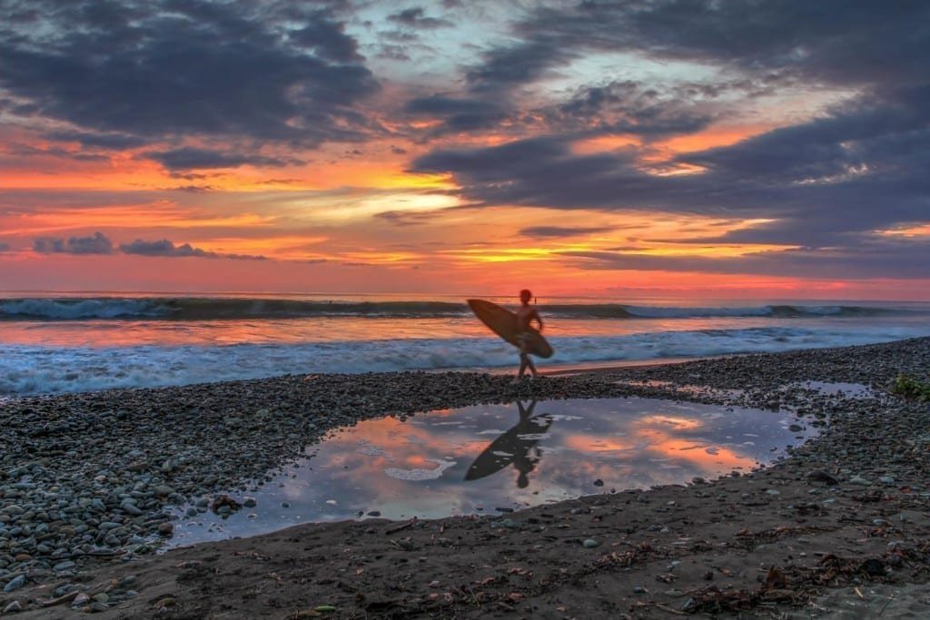 Landed-Travel-Private-Travel-Dominical-Beach-5-Costa-Rica Boy walking down rocky Dominical Beach with a surfboard, backed by vivid fiery sunset over the ocean
