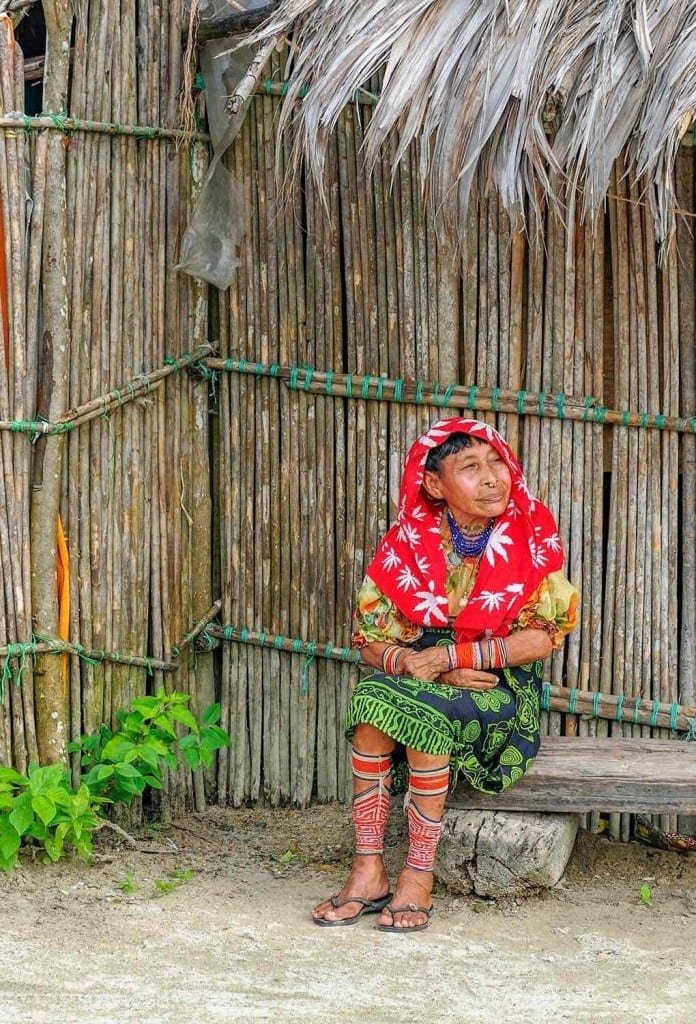 Landed-Travel-Private-Travel-Kuna-Woman-Seated-San-Blas-Panama Indigenous Kuna Woman with a pink shawl and green skirt, sitting on a bench in front of a reed wall in the San Blas Islands