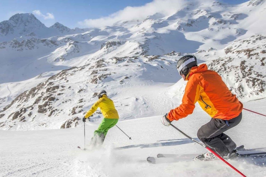 Landed_Travel_Andes_Skiing_3 Couple on their honeymoon in Chile, skiing the powdery slopes of the Andes. Man wearing bright orange jacket & black pants. Woman Wearing Yellow and Green