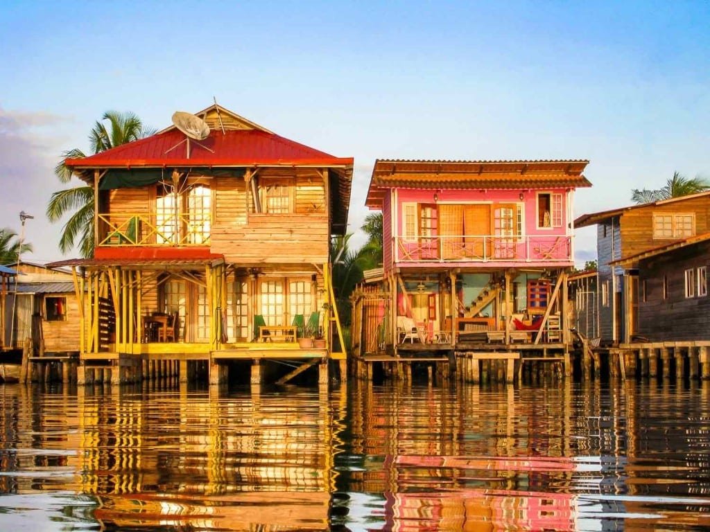 Panama-Private-Custom-Travel-Bocas-del-Toro-Houses PTwo over water bungalos on stilts, one pink with a brown roof, the other brown with a red roof. Over the water of Bocas del Toro