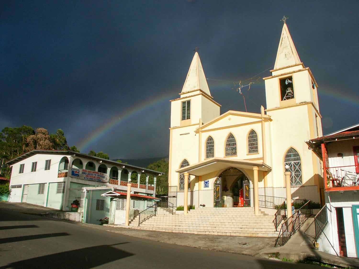 Private-Custom-Travel-Design-Panama-Boquete-Rainbow A beatiful rainbow over a dark sky behind a white twin towered church with pointed rooftops in Boquete, Panama