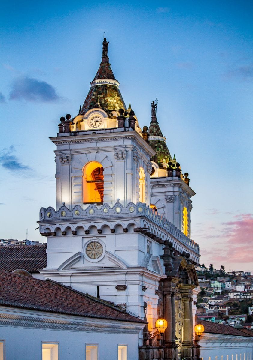 Private-Travel-Custom-Travel-Ecuador-Quito-San-Francisco-Tower Historic White San Francisco Bell Towers, with pointed brown roofs, illuminated at dusk in Quito, Ecuador