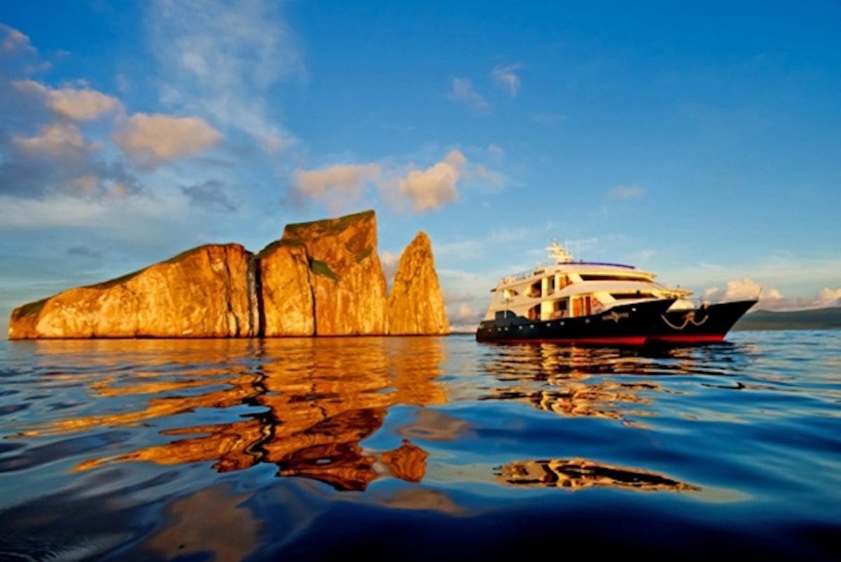 Ocean-Spray-Motor-Catamaran-Charter-Galapagos Blue and White Ocean Spray Luxury Galapagos Catamaran Charter cruising the Galapagos Archipelago near a large orange rock formation