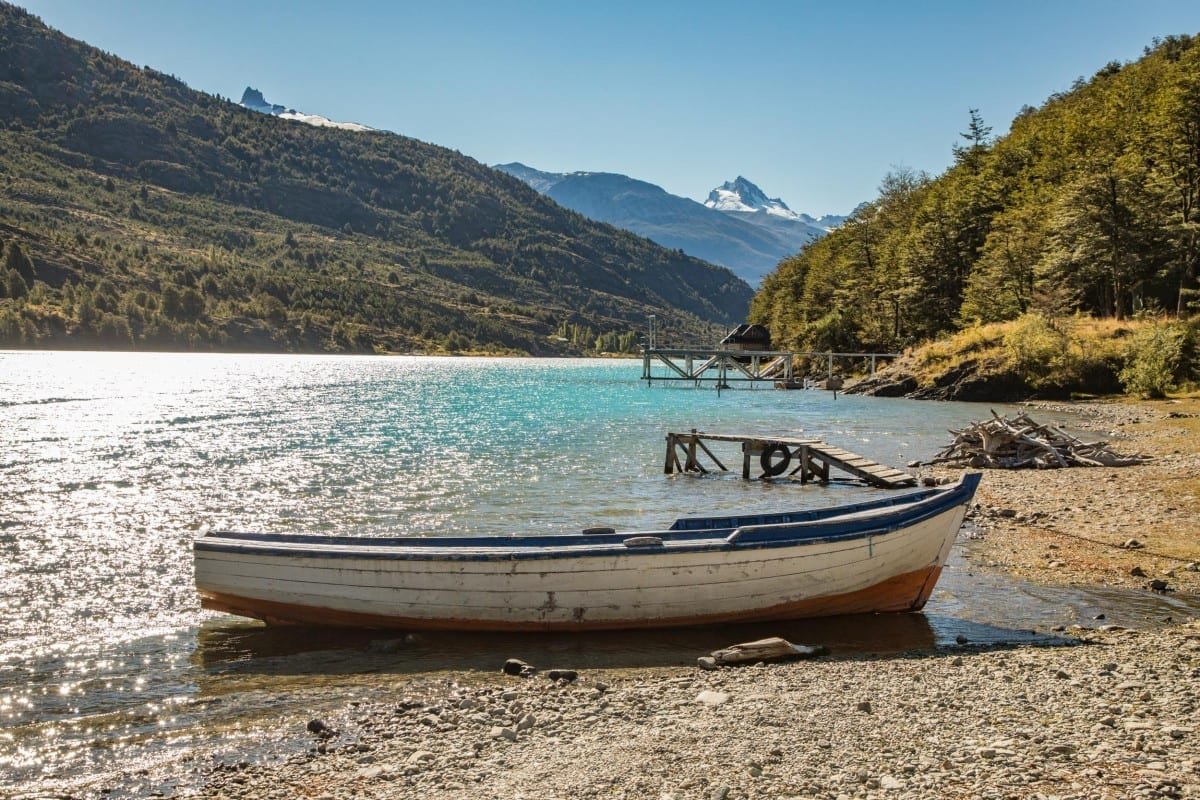 Landed-Travel-Chile-Aysen-9 Small white wooden boat with blue and red accents on the shore of an electric blue lake in Chile off Route 7 in Patagonia