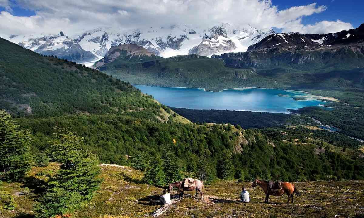 Nibepo-Aike-excursions Three horses graze on a grassy hillside above a bright blue lake and snow-capped mountains in the distance.