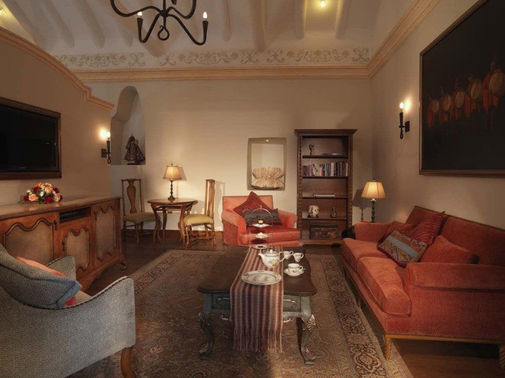 Belmond-Palacio-Nazarenas-One-Bedroom-lounge-Adrian-Houston The lounge in the one bedroom suites at Belmond Palacio Nazarenas feature pink couches, a colorful rug, a tv, paintings and bookshelf.