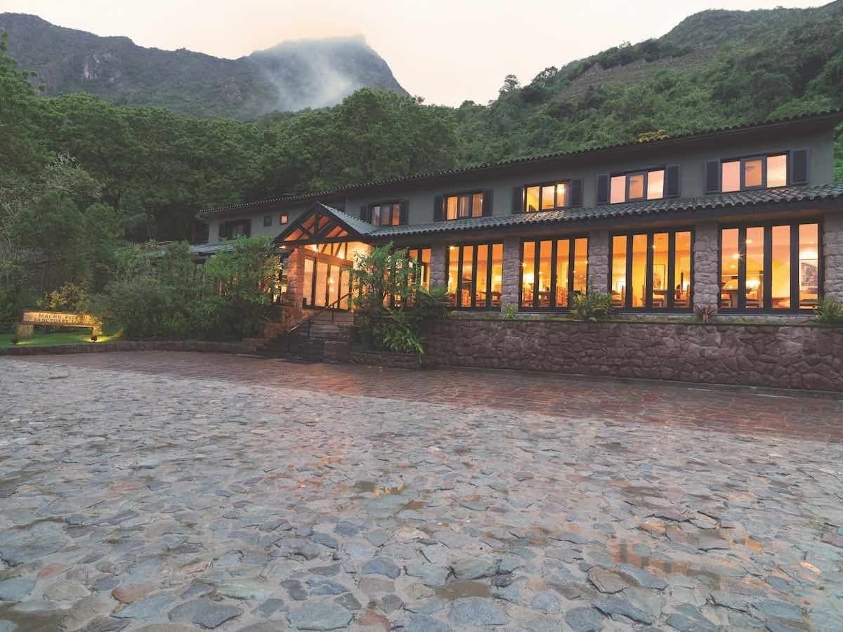Belmond-Sanctuary-exterior The Belmond Sanctuary exterior is a dimly lit lodge with green mountains in the background.