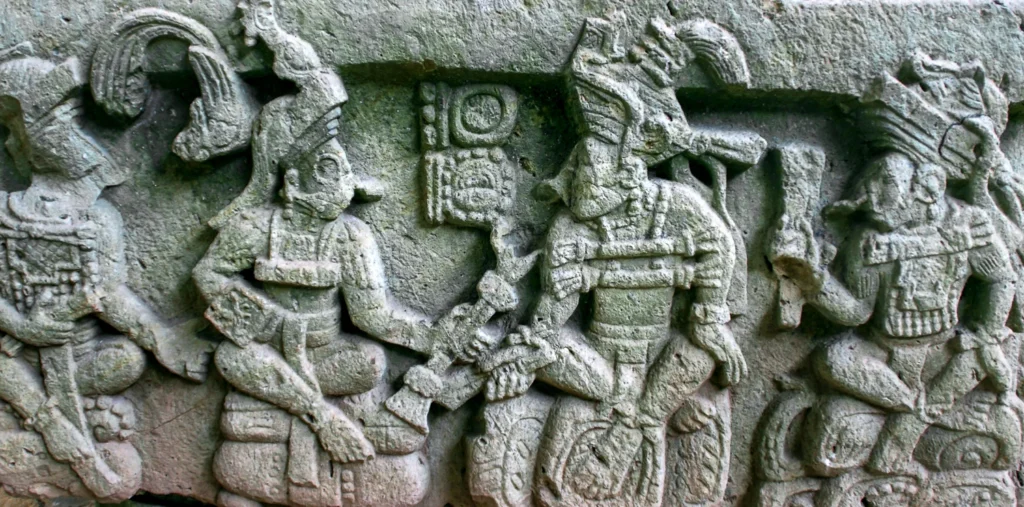 An ancient stone relief carving depicting four seated Mayan figures in elaborate ceremonial headdresses, likely from the archaeological site of Tikal, Guatemala.