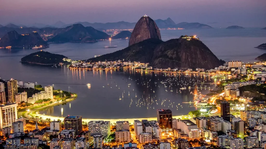 A nighttime twilight view of Rio de Janeiro from above, featuring the illuminated Sugarloaf Mountain in the center, surrounded by a calm bay with boat lights and a glowing city coastline.