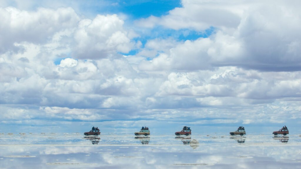 Five all-terrain vehicles driving in a straight line across the flooded salt flats of Salar de Uyuni, Bolivia. The thin layer of water creates a perfect mirror reflection of the vehicles and the massive, fluffy white clouds filling the blue sky above.