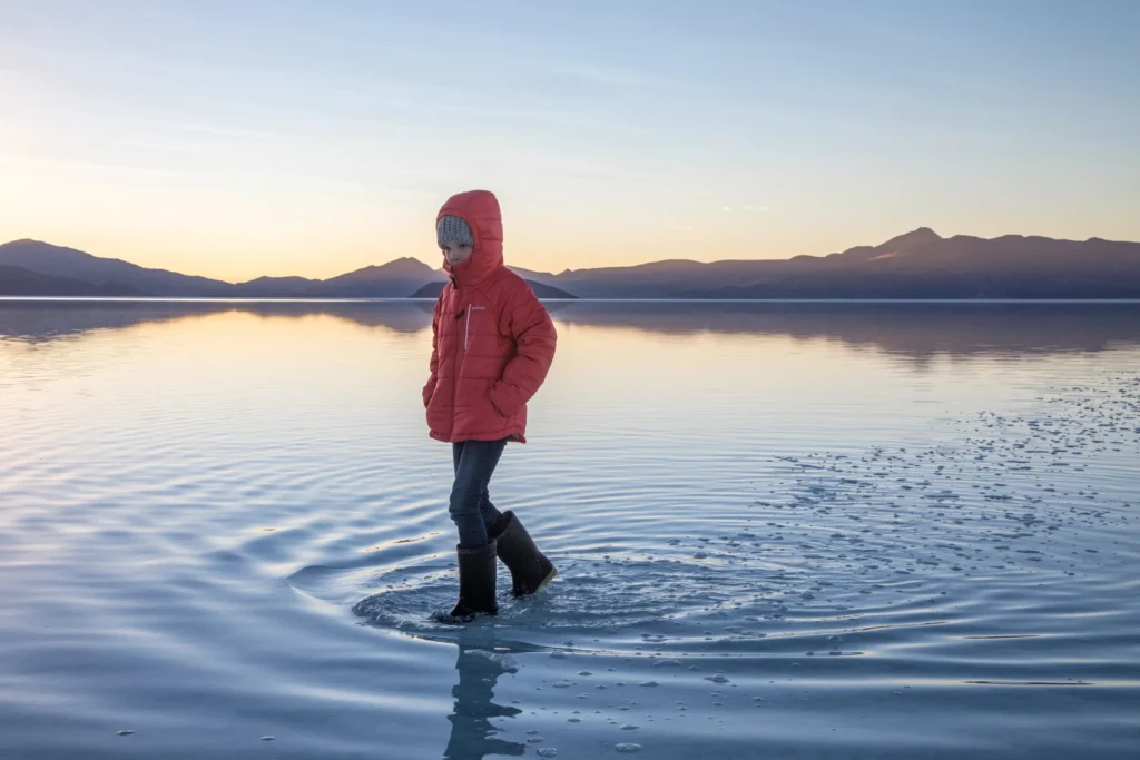Child in a red puffer jacket wading across the mirror-like flooded Salar de Uyuni, Bolivia, at sunset