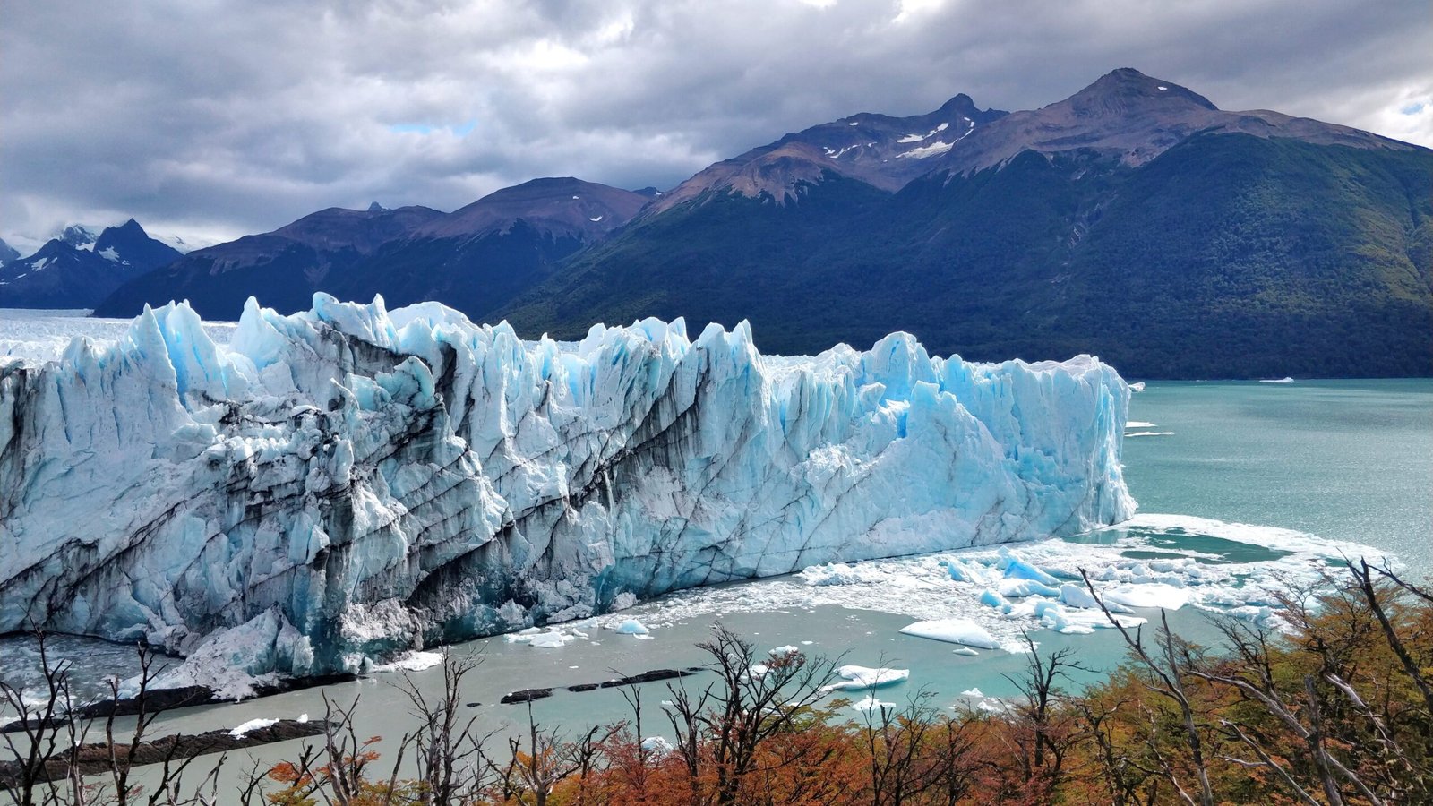 A wide, towering wall of a bright blue and white glacier meets a body of water filled with small icebergs, set against a backdrop of dark, rugged mountains under an overcast sky. In el calafate.