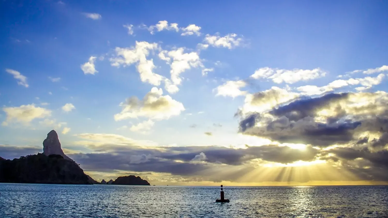 A dramatic oceanic seascape at sunset with bright sunbeams breaking through low, dark clouds over a calm sea. A distinctive, jagged volcanic rock peak stands tall on the left horizon.