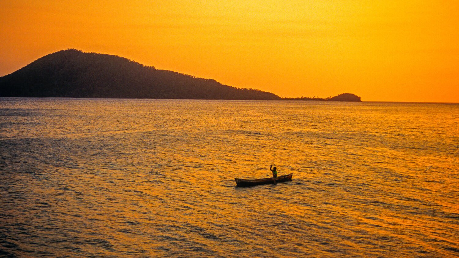 A serene golden sunset over the water in Honduras, featuring a small wooden canoe with a lone person paddling in the calm water. The vibrant orange sky reflects beautifully on the surface of the sea, with a dark, forested island mountain rising in the background.