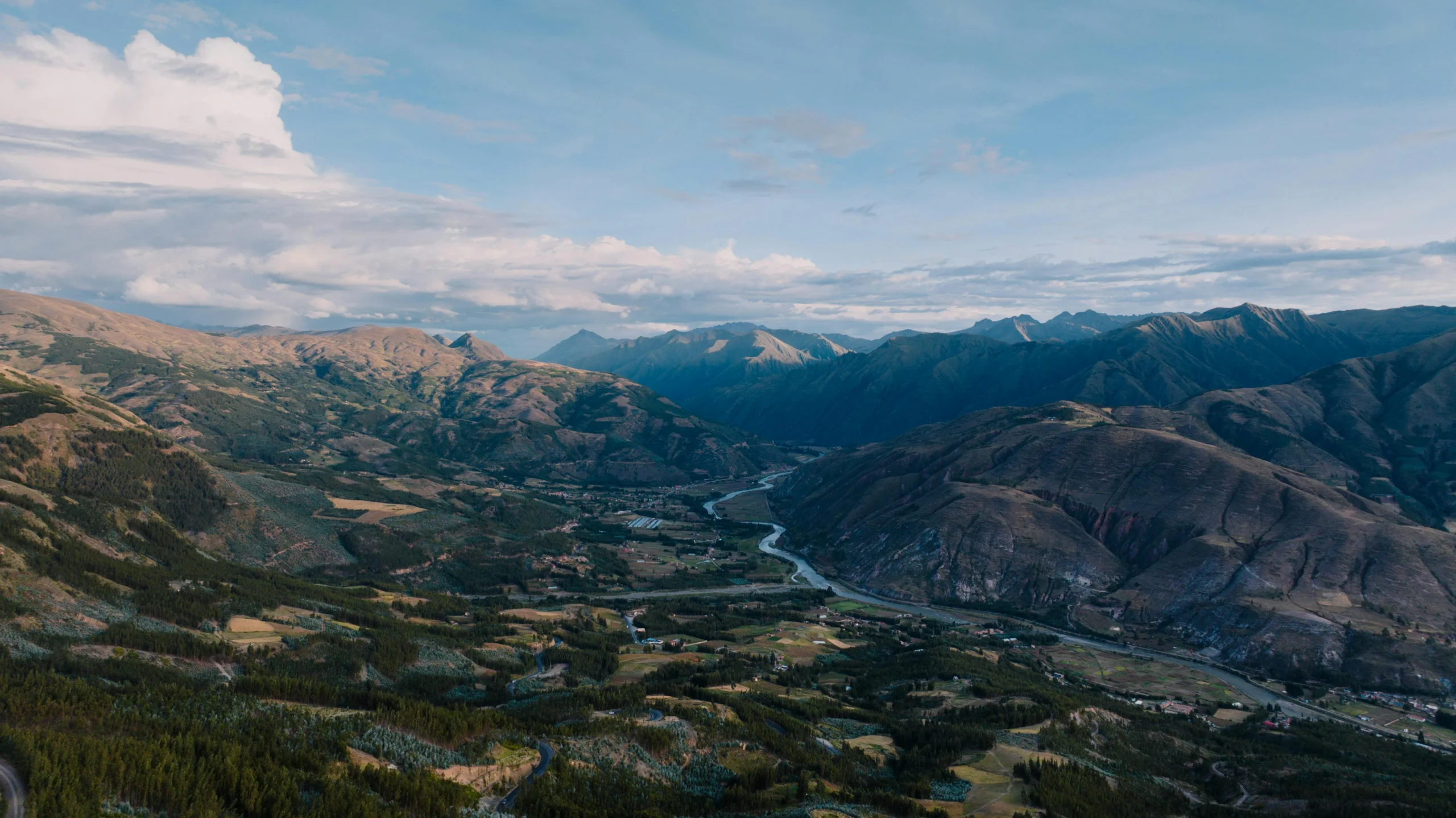 A high-angle panoramic view of the Sacred Valley, showing a winding river cutting through green agricultural fields, flanked by massive, dramatic Andean mountain ranges under a cloudy sky.
