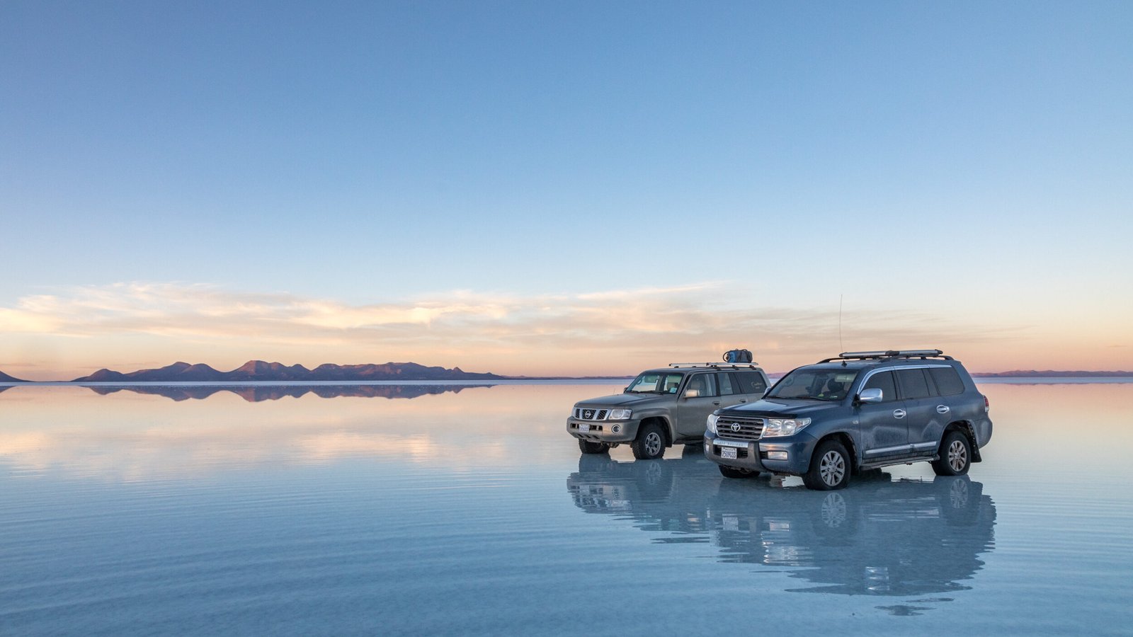 Two off-road vehicles parked on the mirror-like reflective surface of the Salar de Uyuni salt flats in Bolivia during a pale blue and gold sunset.
