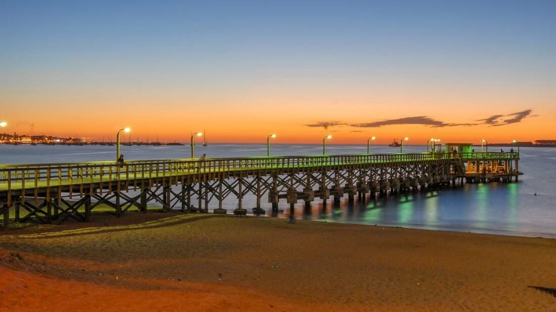 A long wooden pier lit with streetlamps extends over a quiet beach into the ocean at dusk. The sky shows a rich gradient from bright orange at the horizon to deep blue above.