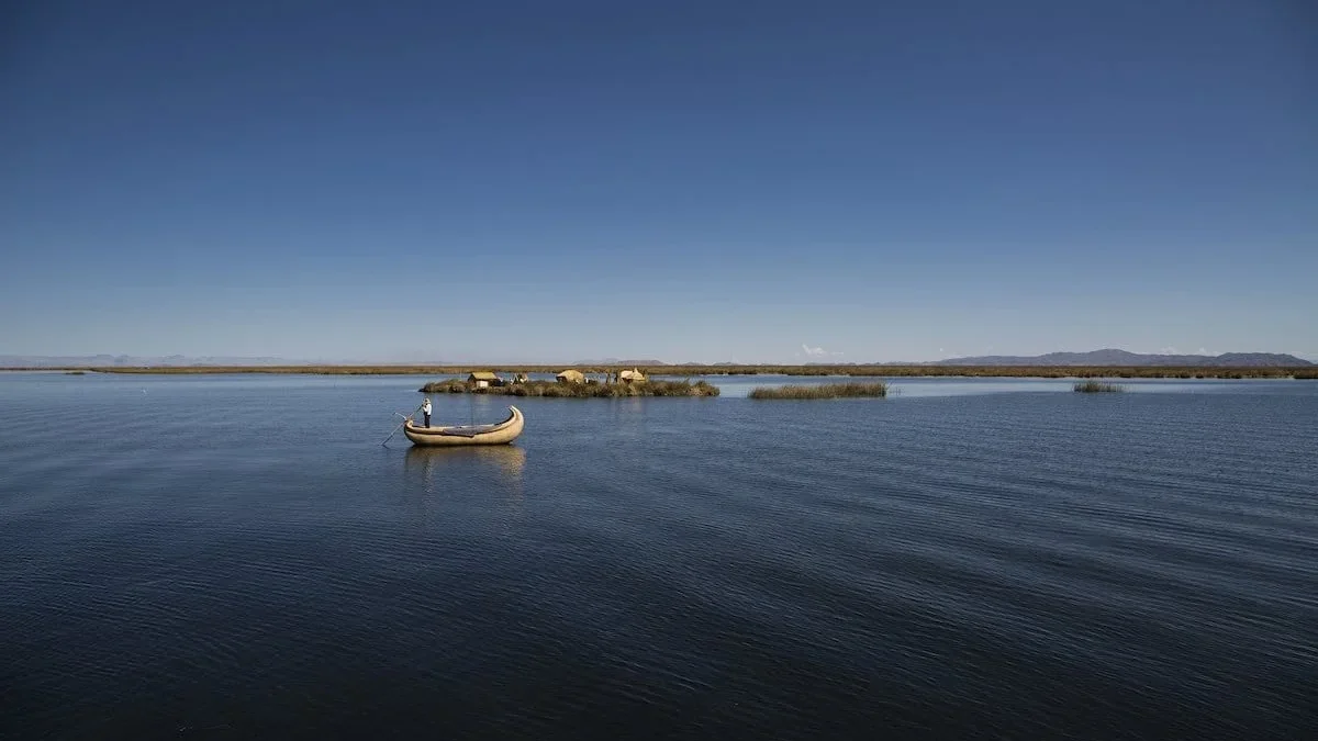 A traditional reed boat floating on the calm, deep blue water of Lake Titicaca near Huatajata, Bolivia. A lone person stands guiding the boat, with a small, floating reed island village and low mountain ridges visible under a vast, clear blue sky.