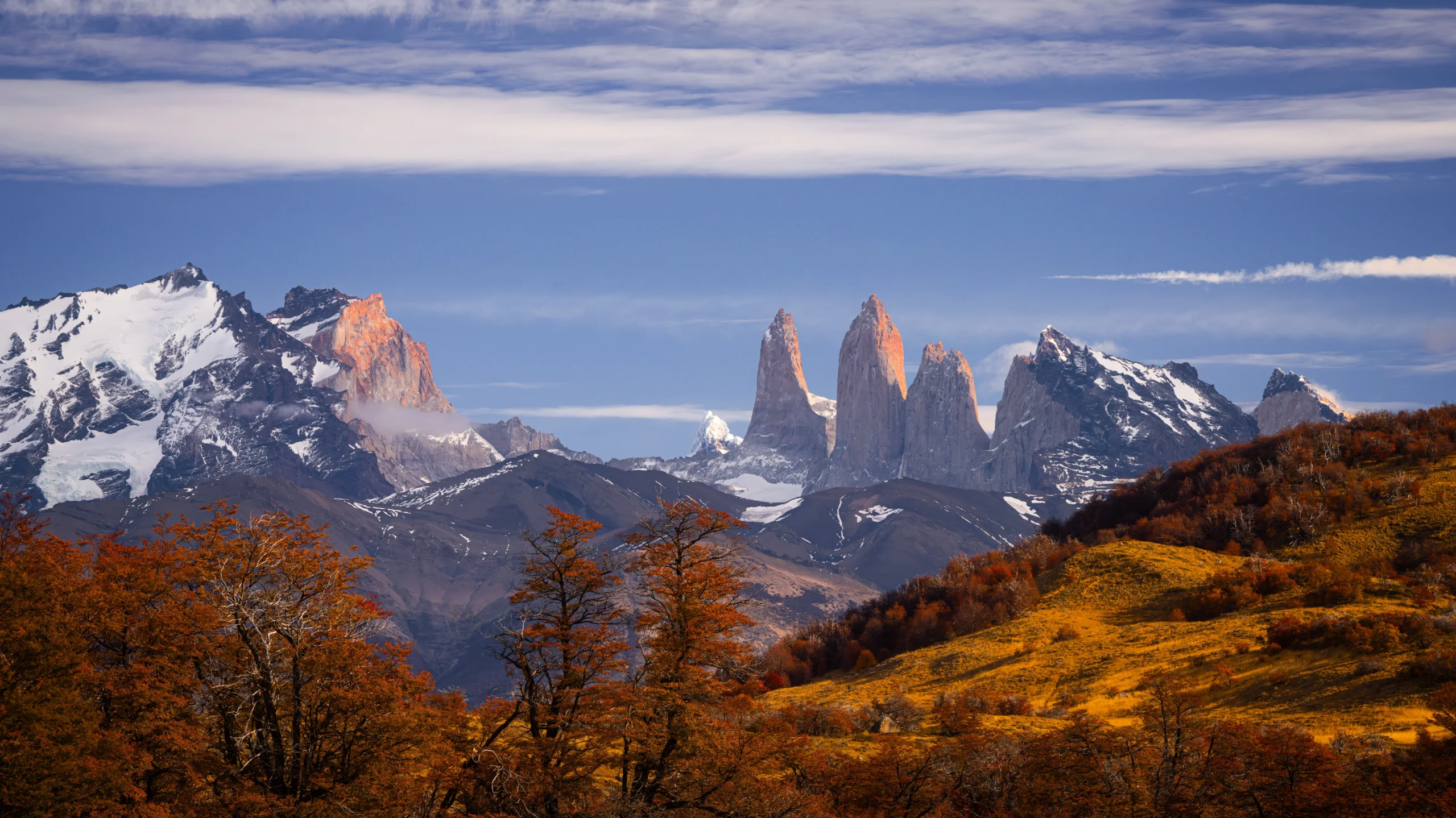 A scenic view of the iconic granite towers of Torres del Paine rising majestically in the background under a blue sky with streaky clouds. The foreground features rolling hills covered in golden and orange autumn foliage, with snow-dusted mountain peaks framing the rugged Patagonian landscape.