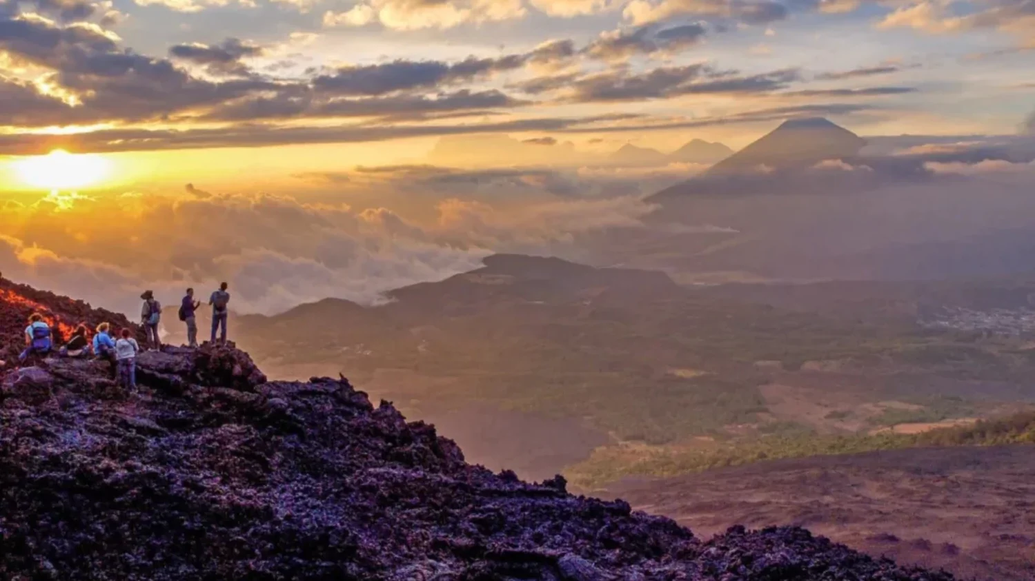 A group of hikers standing on a rugged volcanic slope at sunset, overlooking a vast valley with a distant mountain peak peeking through a layer of clouds