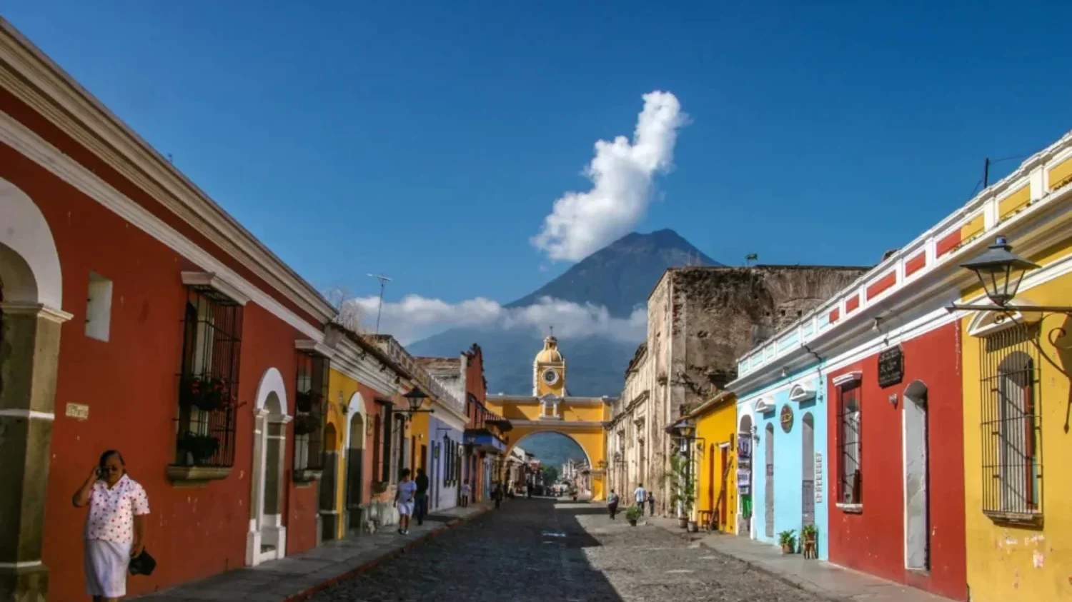 A narrow cobblestone street in Antigua Guatemala, featuring the iconic yellow Santa Catalina Arch and a towering volcano in the background