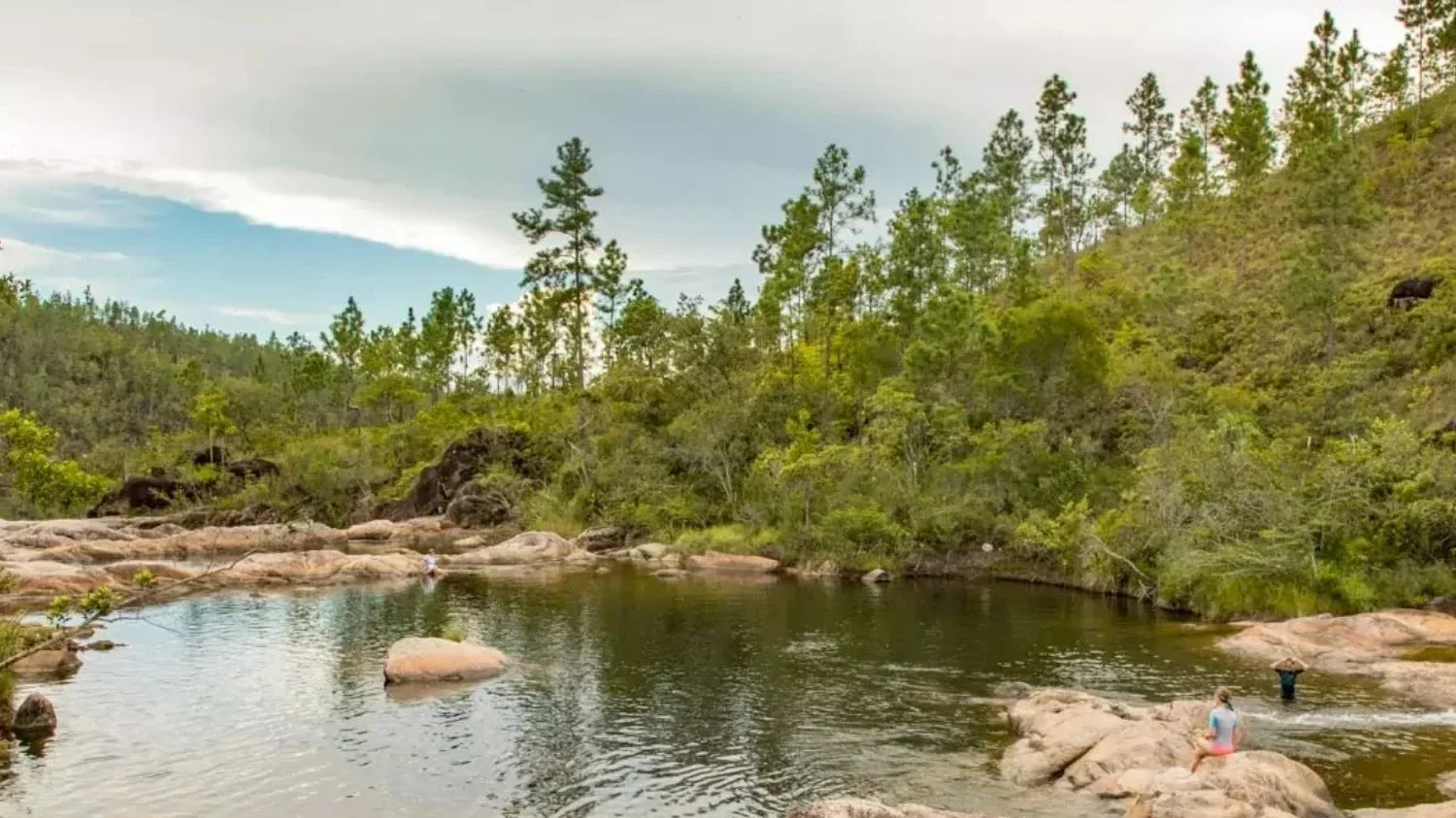 A tranquil, dark-water natural pool surrounded by smooth rock formations and a forested hillside filled with tall pine trees under a soft, overcast sky.
