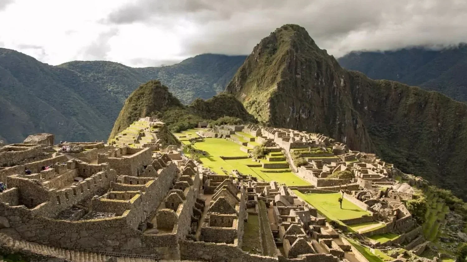 A high-angle, wide view of the ancient Inca citadel of Machu Picchu, Peru, nestled on a green mountain ridge. The stone ruins, terraces, and central green plazas are clearly visible, framed by the towering peak of Huayna Picchu and surrounding misty mountains under an overcast sky.