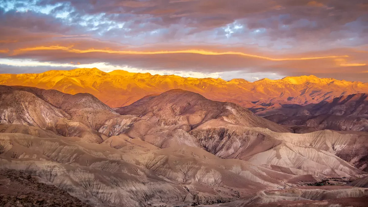 A sweeping panoramic view of the rugged, undulating mountain ridges of the Atacama Desert under a dramatic sunset sky. The setting sun casts a vibrant golden glow across the distant mountain peaks, contrasting with the deep shadows in the dry valleys below.