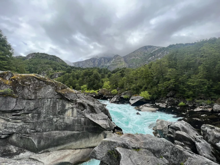 The bright turquoise waters of the Futaleufú River squeeze through rugged, textured gray boulders. Dense green forests line the riverbanks, leading up to towering mountain peaks partially obscured by low-hanging storm clouds.
