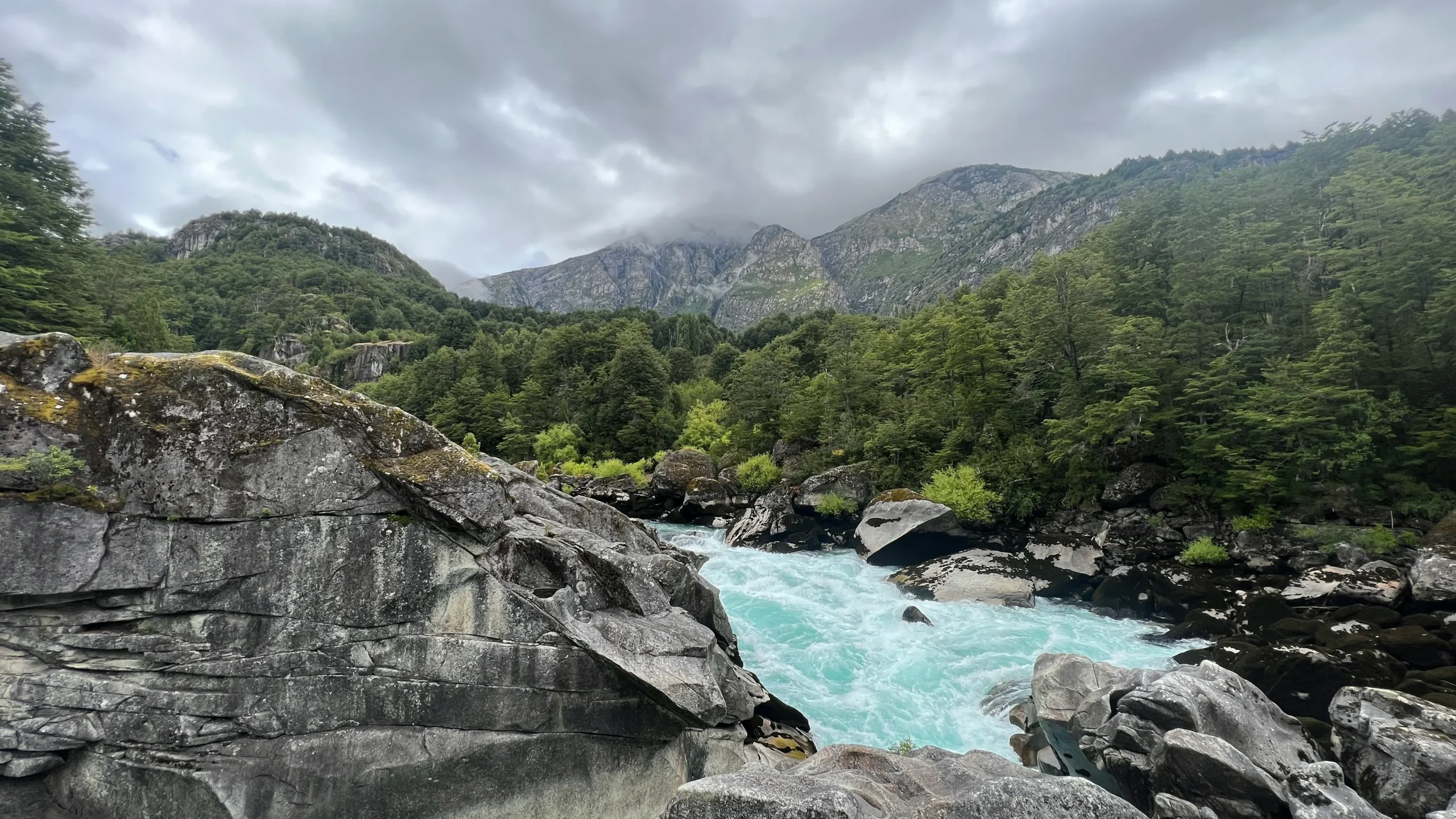 The bright turquoise waters of the Futaleufú River squeeze through rugged, textured gray boulders. Dense green forests line the riverbanks, leading up to towering mountain peaks partially obscured by low-hanging storm clouds.