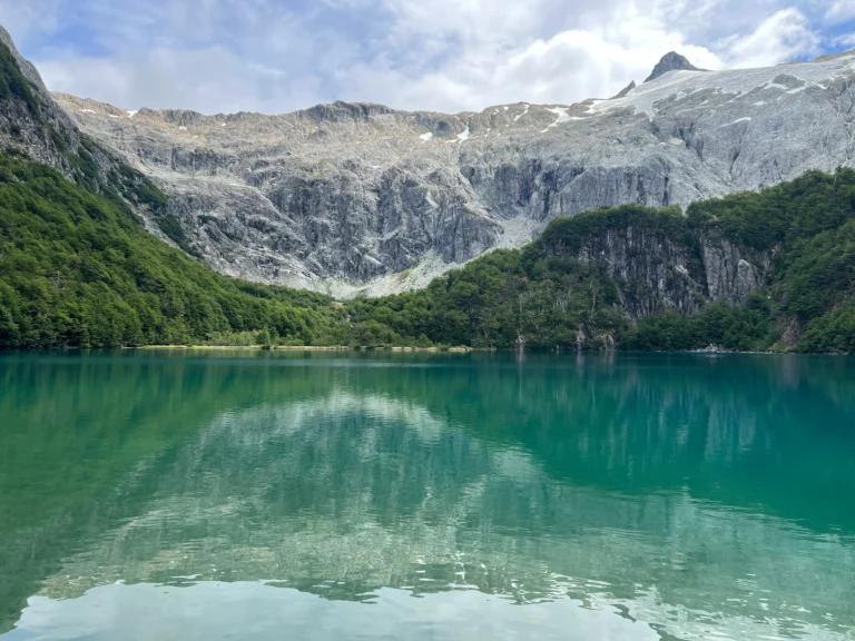A calm, clear emerald-green lake perfectly reflecting a steep, rocky mountain amphitheater and dense green forests under a bright sky.