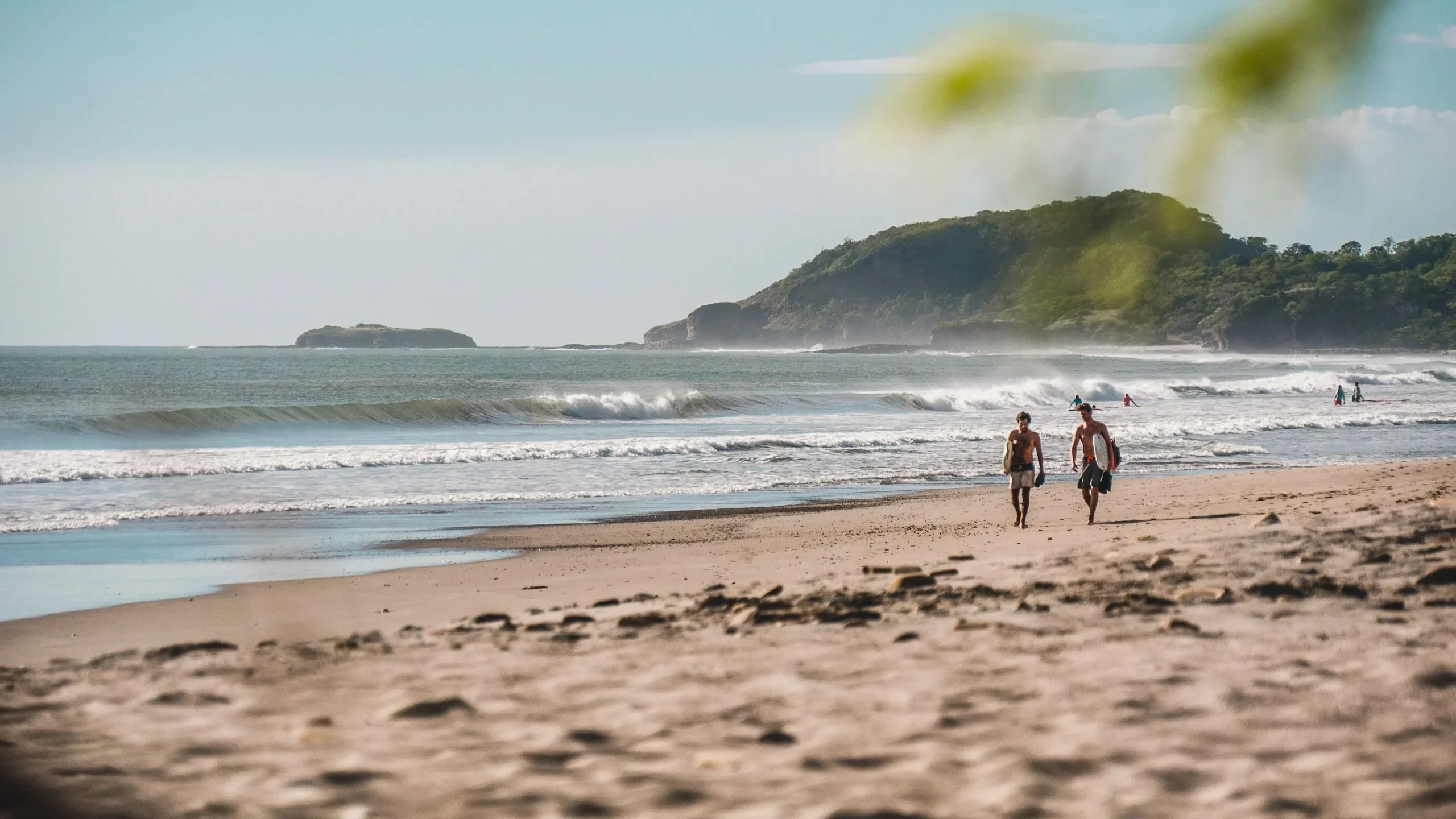 Two surfers walking along a sandy Pacific beach with rolling waves in Nicaragua.