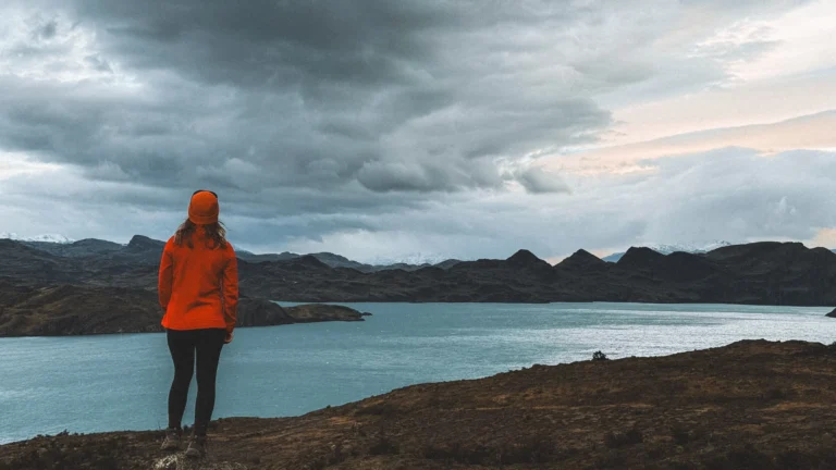 A female hiker in an orange jacket overlooking a massive turquoise glacier lake under dramatic storm clouds in Patagonia, Chile.