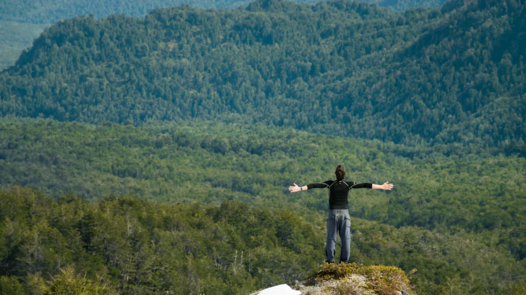 Hiker standing with open arms on a mountain summit overlooking vast, dense green forest valleys in Bariloche, Argentine Patagonia.