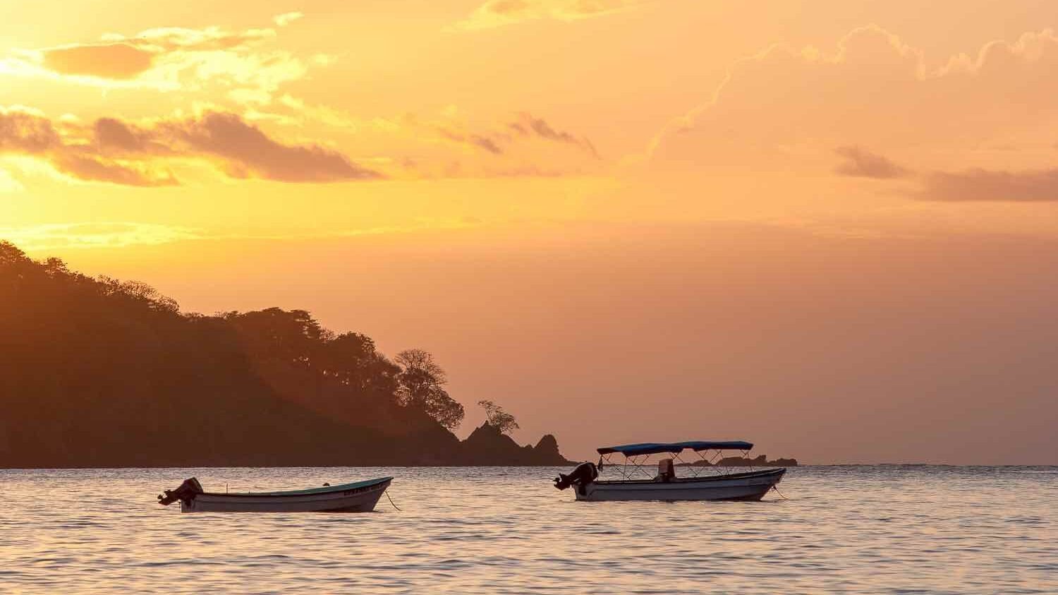 Two small panga boats float on calm coastal waters at sunset, with a dark, forested hillside silhouette in the background under a warm golden sky.