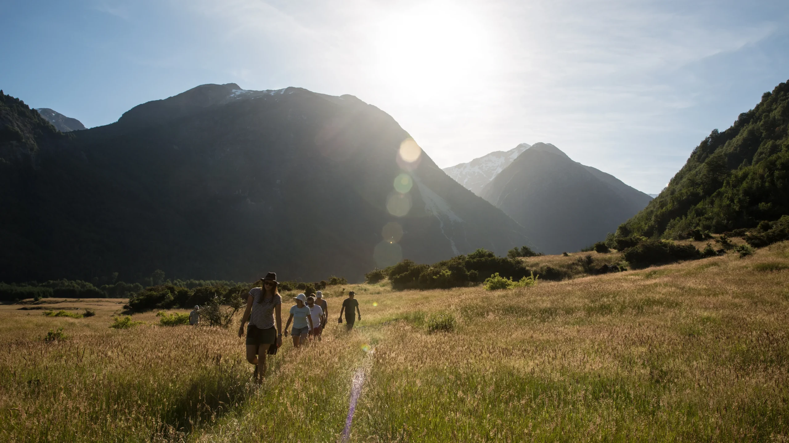 The image captures a group of trekkers silhouetted against the bright sun as they make their way through a golden, sun-drenched field in a mountain valley. The scene evokes a sense of adventure and peace.