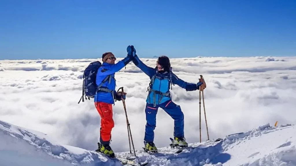 Two hikers celebrate reaching a snowy mountain summit, giving a high-five above a thick layer of clouds under a clear blue sky.